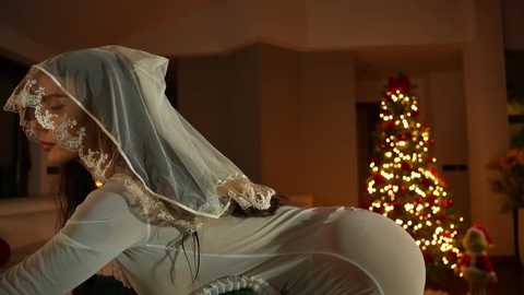 Media: Video of a woman in a sheer, blue wedding veil and dress, bending over a table, with a lit Christmas tree and decorations in the background.