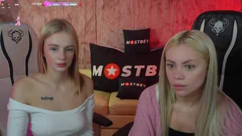 Media: Video of two young women with fair skin and straight blonde hair. One wears a white off-shoulder top, the other a pink cardigan. They sit on a yellow couch, surrounded by black pillows with white skulls.