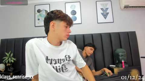 Media: Video of two young men, one in a white T-shirt, the other in a grey hoodie, sitting at a black desk in a minimalist room with abstract art on the wall.