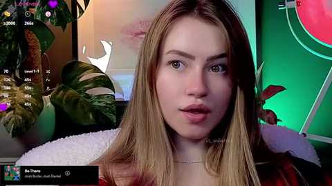 Media: A video of a young, fair-skinned woman with long blonde hair, wearing a red top, and lips slightly parted, indoors with green and purple lighting, plants, and a TV screen showing a clock.