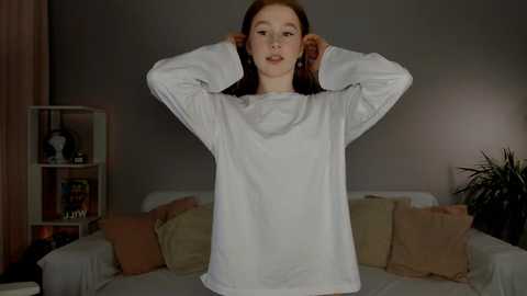 Media: A video of a young woman with fair skin, long red hair, and a white long-sleeved shirt, standing in a dimly-lit living room with a beige sofa, bookshelves, and a potted plant in the background.