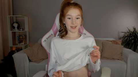 Media: Video of a young Caucasian girl with red hair in a ponytail, wearing a white crop top, lifting it to reveal her belly, in a cozy living room with a beige couch, pink gaming chair, and a bookshelf.