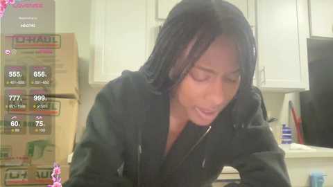 Media: Video of a woman with medium-dark skin, wearing a black hoodie, talking on a phone, in a cluttered kitchen with white cabinets and cardboard boxes.