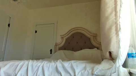 Media: Video of a serene, elegantly decorated bedroom with a plush, white bed, a tufted, cream-colored headboard, sheer curtains, and a partially open door.