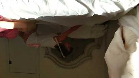 Media: A video of a woman in a white dress peering under a bed with an ornate mirror, reflecting her and a pink bedspread.