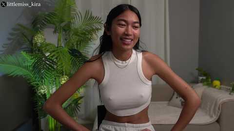 Media: Video of a smiling, slender Asian woman with long black hair, wearing a white crop top and shorts, standing in a modern, indoor living room with a green plant and a beige couch.