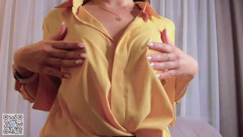 Media: Video of a woman in a yellow blouse, hands cupping her breasts, in a softly lit bedroom with white curtains.