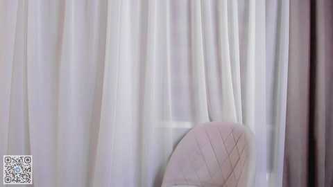 Media: A video of a room with soft white curtains partially drawn, revealing a pale green chair with a quilted pattern. The background is blurred, emphasizing the serene, minimalist decor.