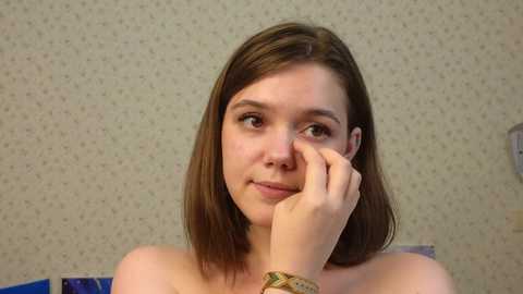 Media: Video of a young Caucasian woman with straight brown hair, fair skin, and light makeup, sitting topless, touching her chin, against a wallpapered beige wall with a subtle pattern.