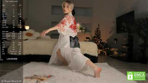 Media: Video of a young, slim woman with short blonde hair, kneeling in a white robe with red floral patterns, in a cozy living room with a Christmas tree, warm lighting, and a bed in the background.