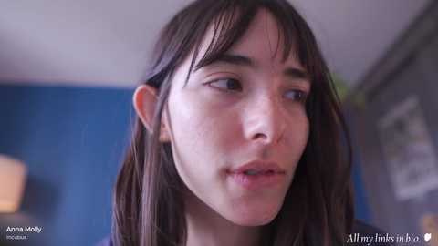 Media: Video of a young, fair-skinned woman with long brown hair and bangs, looking introspective. Background features a blue wall and blurred objects. Text reads \"AllMyLinks in IMDb.\