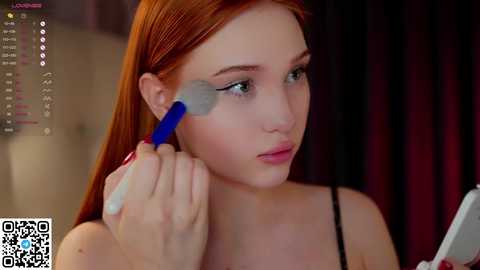 Media: Video of a young woman with long red hair, wearing a black top, using a blue brush to apply makeup on her left cheek. Background features a dark curtain and a blurred headboard.