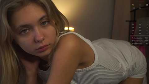 Media: Video of a young, fair-skinned, blonde woman in a white, ribbed tank top and shorts, leaning forward, with a neutral expression, in a dimly lit room with a TV and bookshelf in the background.