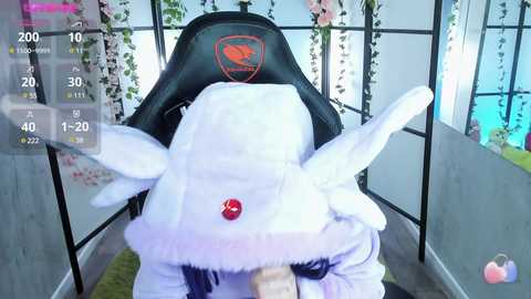 Media: A video of a plush toy resembling a white dragon with red eyes, seated in a black gaming chair, surrounded by pink and white floral decor, with a virtual gaming overlay displaying stats and controls.