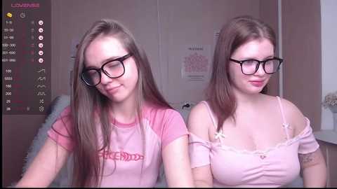 Media: Video of two young women with long hair, wearing glasses and pink shirts, sitting side by side in a bedroom with a calendar and posters on the wall.