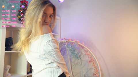 Media: Video of a blonde woman in a white striped shirt, facing away, standing in a room with a pink and blue globe, a Christmas wreath, and a white shelf.