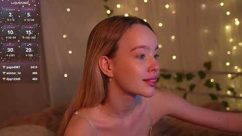 Media: Video of a young woman with straight, light brown hair, wearing a white tank top, looking to the side in a cozy, dimly lit room with string lights and a digital clock showing 3:37 AM.