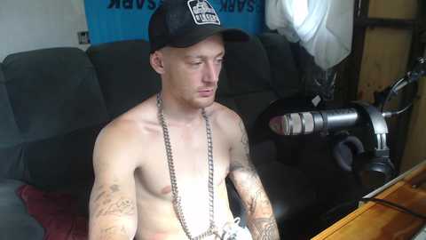 Media: Video of a shirtless, tattooed, fair-skinned, young man with a black baseball cap, wearing a silver chain necklace, sitting in a dimly lit room with a grey couch and studio lighting equipment.