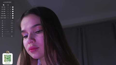 Media: Video of a young woman with long, straight dark brown hair and light skin, wearing a white top, captured in a dimly lit room with gray curtains.