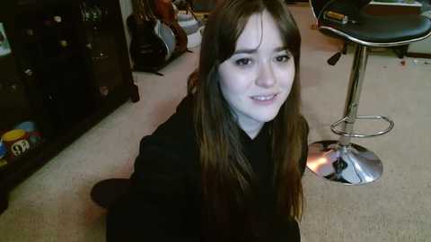 Media: Video of a young woman with long brown hair, fair skin, and a casual black outfit, kneeling on a beige carpet in a cluttered room with dark furniture, guitars, and a stool.