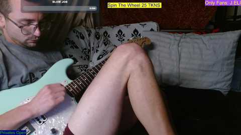 Media: Video of a man with glasses, fair skin, and short brown hair, playing a white electric guitar, sitting on a patterned couch, in a dimly lit room.