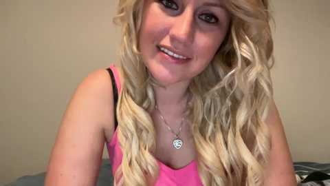 Media: Video of a smiling, fair-skinned young woman with long, wavy blonde hair, wearing a pink tank top and a silver necklace with a heart pendant. She has a friendly, open expression.