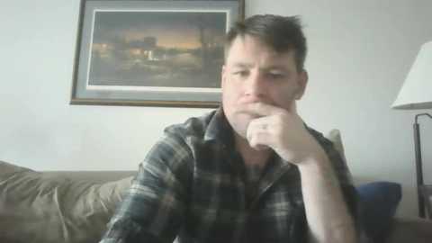 Media: A video of a middle-aged man with short, dark hair, wearing a plaid shirt, resting his chin on his hand, in a living room with a framed painting on the wall and a beige couch.