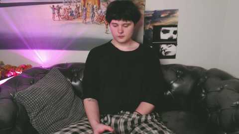 Media: Video of a young man with short black hair, wearing a black shirt and plaid pajama pants, sitting on a gray, tufted leather couch in a dimly lit room.