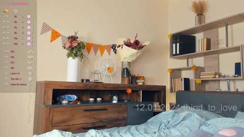 Media: Video of a cozy bedroom with wooden furniture, a calendar, orange and white bunting, flowers, books, and a blue bedspread.