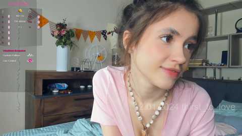 Media: Video of a young woman with light skin and dark hair, wearing a pink shirt and beaded necklace, sitting on a bed in a cozy bedroom.