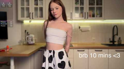 Media: A video of a young, fair-skinned girl with long brown hair in a white, polka-dot crop top and skirt, standing in a modern kitchen with white cabinets and a wooden countertop.