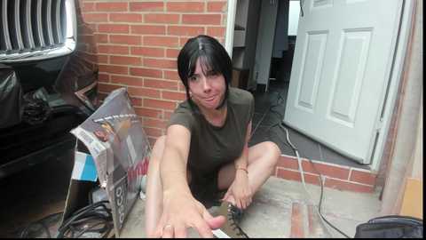 Video of a woman with short black hair, sitting on the ground in front of a brick wall and white door, wearing a green t-shirt, surrounded by clutter.