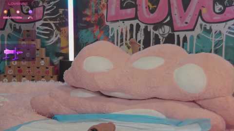 Media: A video of a plush, pink, polka-dotted teddy bear with a soft, fluffy texture, lying on a bed with a white sheet. The background features graffiti art with the word \"LOVE\" in large, colorful letters.