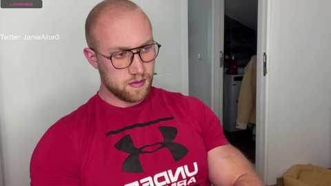 Media: Video of a muscular, bald, fair-skinned man with a beard and glasses, wearing a red Under Armour t-shirt, standing in a dimly-lit room with white walls and a partially open door.