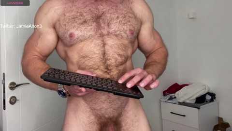 Media: Video of a muscular, nude, hairy-chested man holding a keyboard in a bedroom. He has a lean build, fair skin, and a small penis. Background includes a white door, dresser, and scattered clothes.