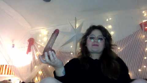 Media: A video shows a young woman with long brown hair and fair skin, wearing black clothing, holding a large, realistic-looking dildo. The background features a festive, cozy room with string lights and a star-shaped decoration.