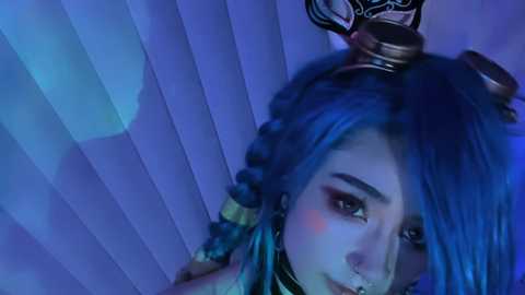 Media: Video of a young woman with blue hair and steampunk goggles, wearing a mask, in a dimly lit, blue-lit room with vertical blinds.