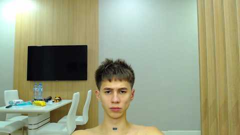 Media: Video of a young man with short, spiky dark hair, looking serious, in a modern, minimalist room with light wood paneling, white chairs, and a flat-screen TV.