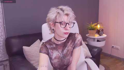 Media: A video of a pale-skinned, androgynous person with short, platinum-blonde hair, wearing black-rimmed glasses, a black choker, and a tie-dye shirt, sitting on a black leather couch in a dimly-lit room.