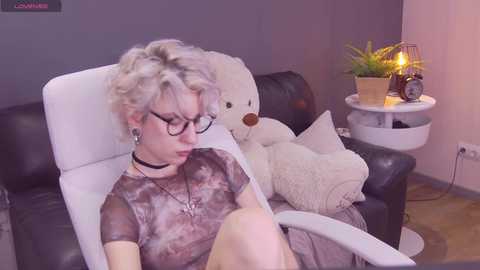 Media: Video of a pale-skinned woman with short, platinum-blonde hair, wearing black-rimmed glasses, a black choker, and a tie-dye t-shirt, seated in a black leather chair. Background features a plush white teddy bear, a small white side table with a plant and a lamp, and a dark wall.