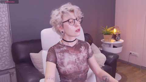 Media: Video of a pale-skinned, short-haired woman with glasses, wearing a tie-dye shirt, black choker, and tattoos, sitting on a black leather chair in a modern, dimly-lit room with a small plant on a white side table.