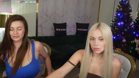 Media: Video of two young women: one with long brown hair in a blue dress, and another with long blonde hair in a strapless top. Background includes a Christmas tree and two black pillows with \"MST\" text.