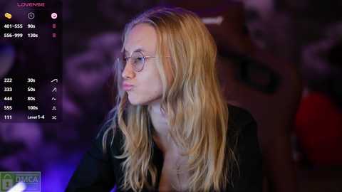 Media: Video of a young woman with long, wavy blonde hair, wearing round glasses and a black top, gazing pensively. Background features dimly lit, blurred elements.