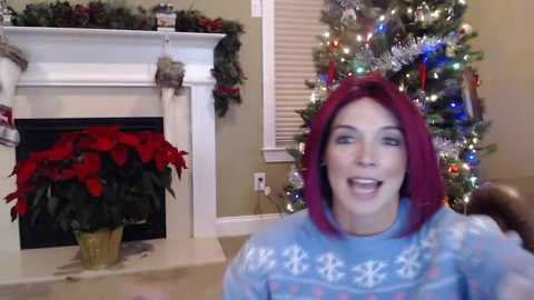 Media: Video of a woman with bright red hair, wearing a blue Christmas sweater, smiling widely, in a festive living room with a decorated Christmas tree and poinsettia.