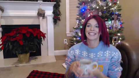 Media: Video of a smiling woman with red hair in a blue holiday sweater, holding a coffee mug, in a festive living room with a Christmas tree and potted poinsettia.