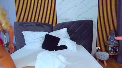 Media: Video of a modern bedroom with a grey upholstered headboard, white bedding, and a black pillow, against a marble and wood-paneled wall. A blonde woman in red lingerie is partially visible on the left.