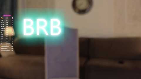 Video of a dimly-lit living room with a green neon \"BRB\" sign on a wall, a clock, and a couch in the background.