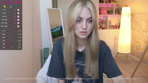 Media: Video of a young, fair-skinned woman with long blonde hair, wearing a dark T-shirt, sitting indoors. Background shows a cozy, well-lit room with a mirror, shelves, and colorful decor.