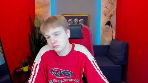 Media: Video of a young, fair-skinned, blonde male in a red sweatshirt with white sleeves, sitting in a red chair with a black cushion, surrounded by red and blue walls.