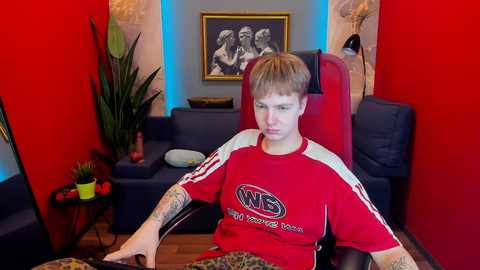 Media: Video of a young person with short, light brown hair, wearing a red \"Nike\" T-shirt, seated on a red gaming chair in a brightly lit, red-walled room with a potted plant, framed artwork, and a dark blue sofa.